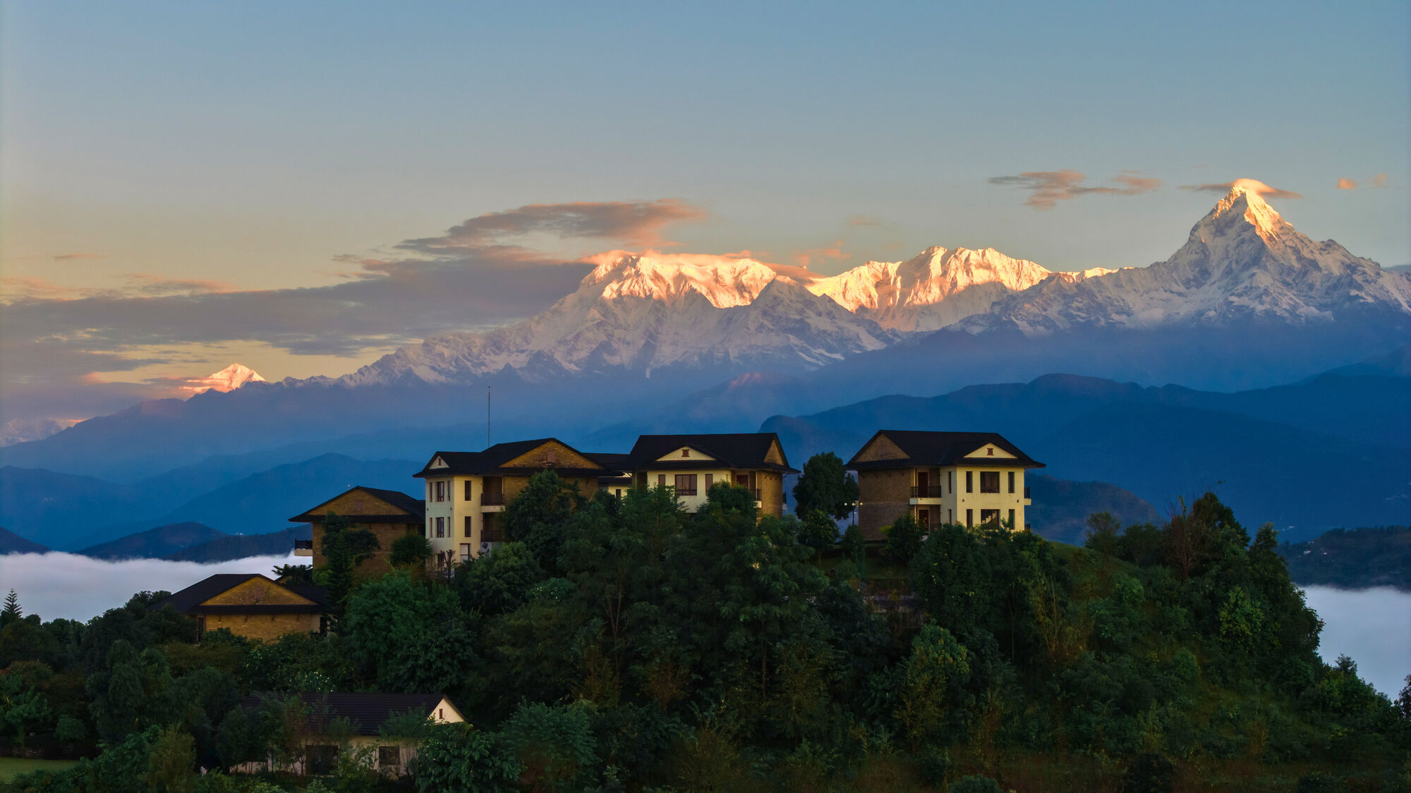 Rupakot Resort villas on a green hilltop, with cloud below and the snow-covered Annapurna range behind